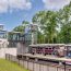 Photo of the MBTA East Taunton commuter rail station ovrelooking the tracks on a sunny day. One train is stationary and another is in motion. There are people on the boarding platform including a conductor and bicyclist.