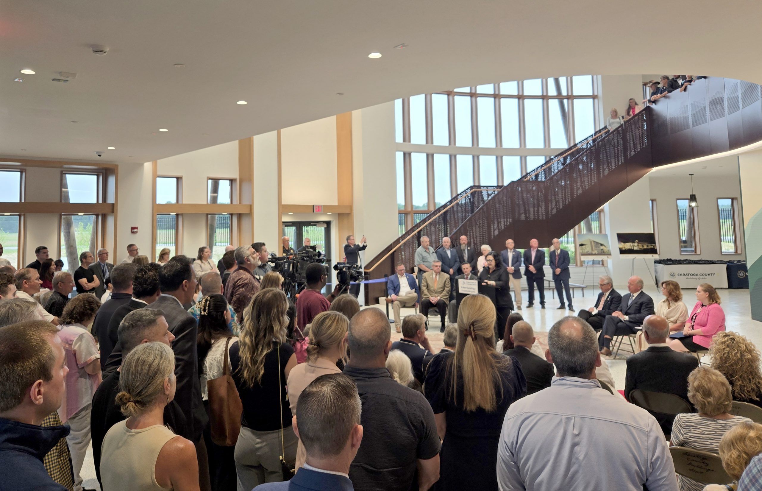Photo of people standing in the Saratoga County Airport atrium listening to officials speak at a podium. The atrium is a high-ceilinged room with a wall of windows in the background. A large staircase in the behind the speakers leads to the upper level.