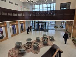 Photo of the Saratoga County Airport atrium. The atrium is a high-ceilinged room with a wall of windows in the background. This view is looking toward the main entrance. It was taken from the second level looking down at chairs and tables on the first floor.