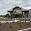 Photo of the exterior and landside entrance of the Saratoga County Airport. There is a round landscaped area in front with a bronze horse and jockey statue in its center. The front facade of the building is primarily glass with CLT timber supports.