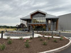 Photo of the exterior and landside entrance of the Saratoga County Airport. There is a round landscaped area in front with a bronze horse and jockey statue in its center. The front facade of the building is primarily glass with CLT timber supports.