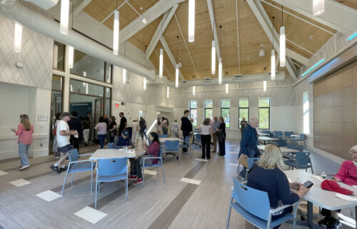 Photo of a large room with an expansive two-story wood ceiling. Multiple lights hang down over tables and chairs. People are gathered and talking in standing groups and at tables for the opening celebration.