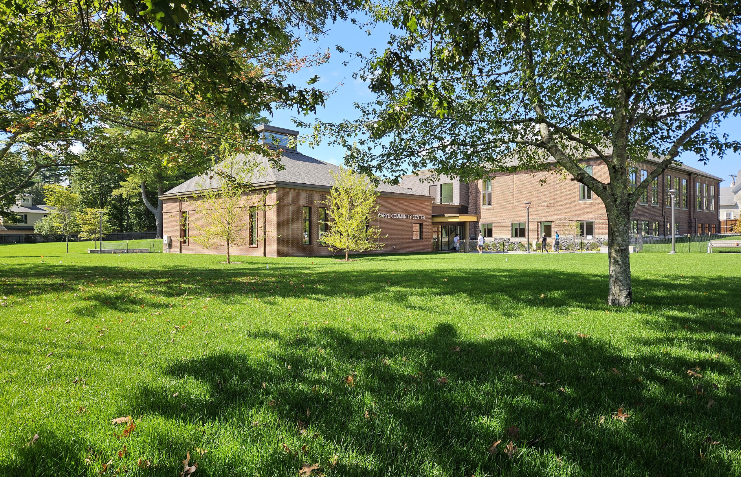 Exterior photo of Caryl Community Center showing the new pavilion and historic school sections. The exterior is brick and set back on a green lawn surrounded by trees.
